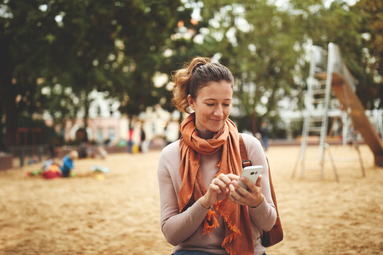 Frau tippt auf ihrem Smartphone auf einem Spielplatz.